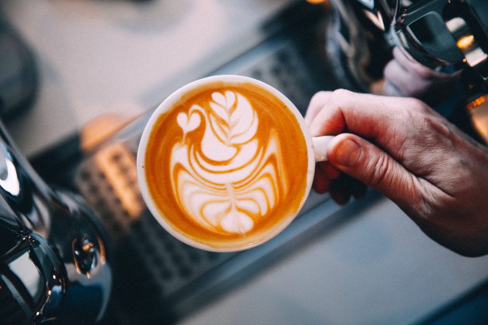 A hand lifting a white cup of coffee with a leaf poured into the foam